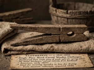 Molletta antica in legno scuro intagliata a mano da un singolo pezzo, appoggiata su tela di iuta con cartellino museale Clothespin 1810-1825, atmosfera seppia